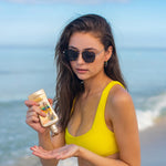 Woman in yellow bikini and sunglasses applying sunscreen on a beach