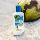 A bottle of sunscreen lotion standing on sand with a blurred coconut in the background. The bottle is white with a blue flip-top cap and a tropical palm tree label showing blue sky and ocean.