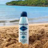 Panama Jack sunscreen spray can on a sandy beach with ocean and trees in the background