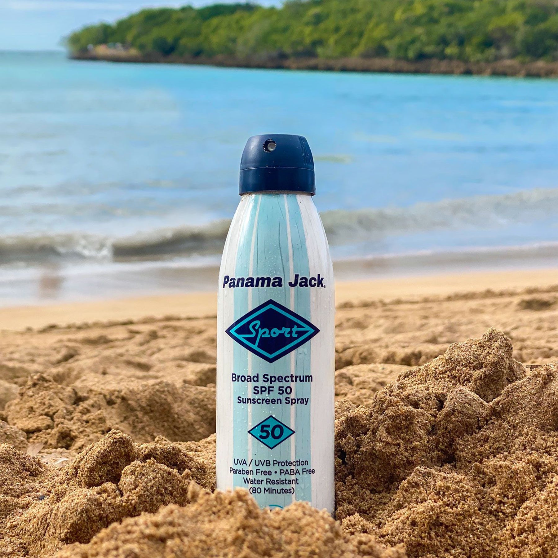 Panama Jack sunscreen spray can on a sandy beach with ocean and trees in the background