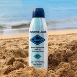 Panama Jack sunscreen spray bottle on a sandy beach with ocean in the background