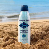 Panama Jack sunscreen spray bottle on a sandy beach with ocean in the background